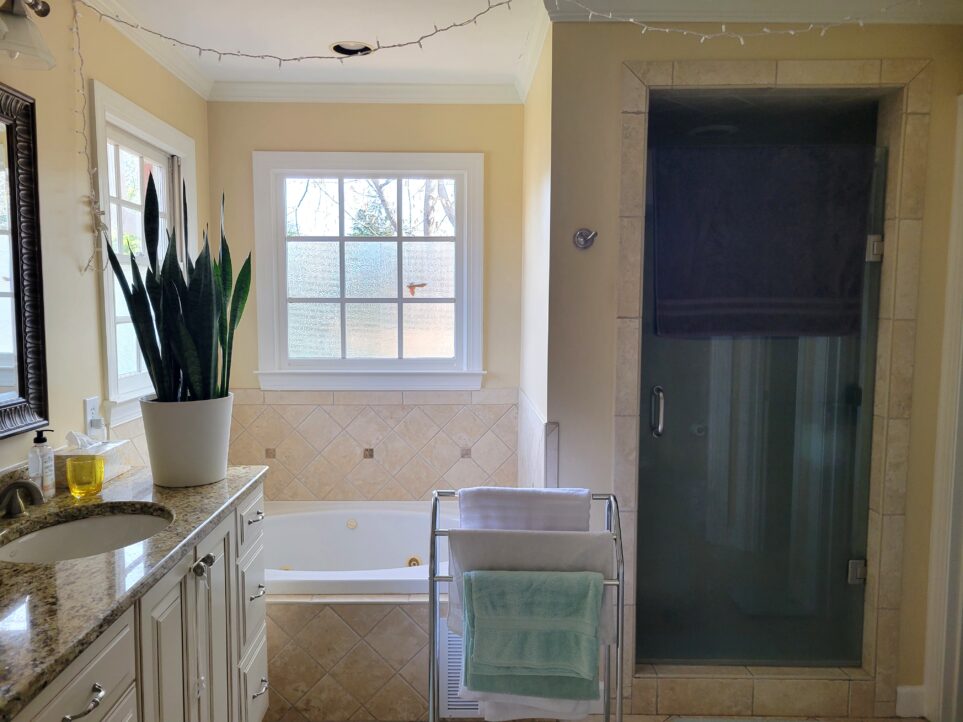 View of primary bathroom from door leading off of primary bedroom. Shower is straight ahead to right (with a free-standing towel rack positioned just to the left of its door, in front of the tub), and beyond it to right (out of sight) are two primary walk-in closets and a toilet stall immediately to the right of the door. Ahead in left corner is a garden tub below two windows. Along wall to immediate left is double sink countertop and vanity (partially out of sight) with snake plant positioned at end facing windows.