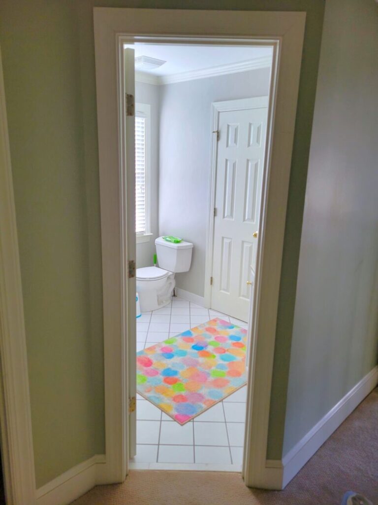 View of bathroom near children's bedroom, standing outside of it in the hallway near the office and looking through open door. Partially in view against far war are toilet and window. The door that cuts through to the children's bedroom closet is to the right of the toilet. A colourful rug with circles in various bright colours lies on the white tile floor in the center of the bathroom.