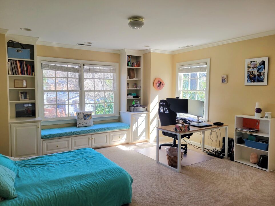 Office viewed from doorway, facing the back corner by the south-facing window and desk. To the left is a floor bed with blue comforter. To the right against back wall is a small white bookcase with some paperwork and other items on the shelves, a tiny lamp on top, and a salt lamp on the bottom shelf. To its left is an L-shaped desk with work equipment set up - computer monitor, laptop, swivel chair, etc. and the south-facing window in the corner. A few framed photos are in view along the wall. To the left of the window, along next wall, is a built-in double window seat with a cushion covered in a blue sheet, and a throw pillow in front of the windows. Flanking both sides of the window seat are built-in bookshelves, storing various textbooks and other items. Sunbeams pour in through the south-facing window