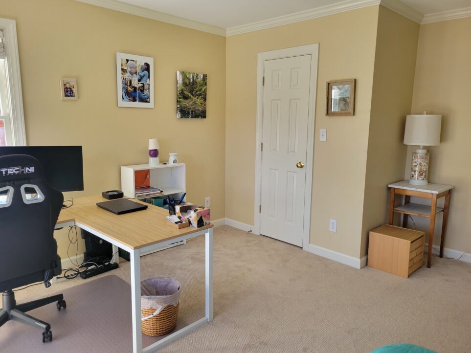 Office viewed from bed, facing corner with closed closet door. In view on the right is a small nook with with an end table in the corner and a lamp on top with seashells in the base. Just next to it on the floor is a small wooden cube full of drawers of children's learning/building pieces. Some framed photos hang on the wall near the closet door and toward the south-facing window, which mostly out of view. In view on the left (in front of the mostly-unseen window) is an L-shaped desk with a swivel chair and computer monitor. To the right of the desk along the south wall is a small white bookcase with some paperwork and a tiny lamp on top.