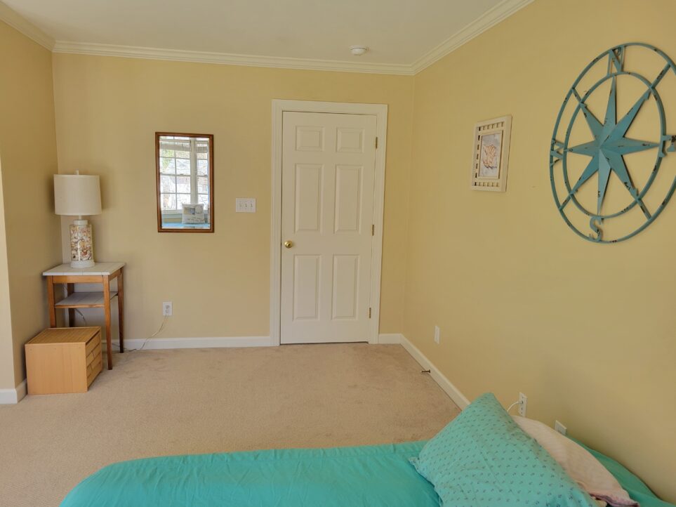 Office viewed from the side of the floor bed, partially seen below and covered in blue quilt. Straight ahead is closed door (leading out into the hallway), to the left of which is a small nook with an end table in the corner and a lamp on top with seashells in the base. To the right a mirror hangs on the wall between the lamp/table and door. A small wooden cube full of drawers of children's learning/building pieces sits next to the end table within the nook. To the right and above the bed hangs a large blue metal compass rose and, further along the wall toward the corner by the door, a framed seashell painting.