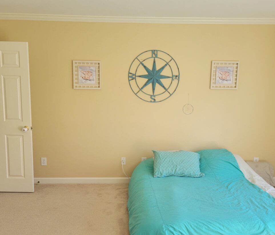 Photo of office, viewing floor bed with blue quilt against pale yellow wall against which hangs two framed seashell paintings and a large blue metal compass rose in between, with a small white/clear dreamcatcher above the bed. Open door (from hallway) is partially seen to the left