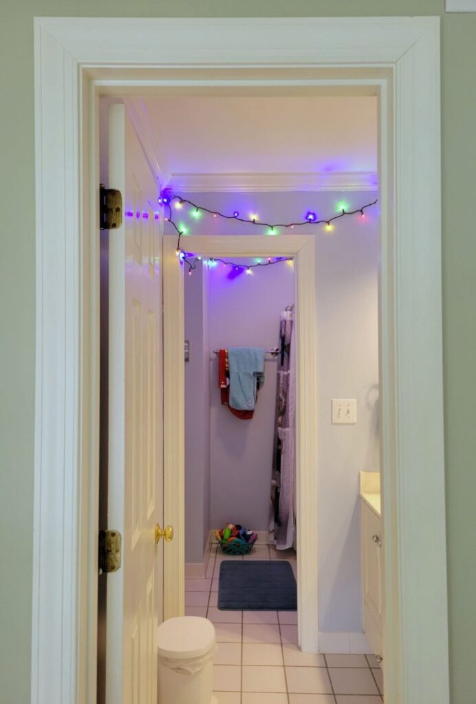 View of hallway bathroom upstairs, standing outside of the door in the hallway and viewing most of the door frame and straight back to the wall by the shower tub. A lit strand of multi-coloured string lights is seen strung across the width of the ceiling in the sink area as well as further back along the ceiling in the toilet/tub area.