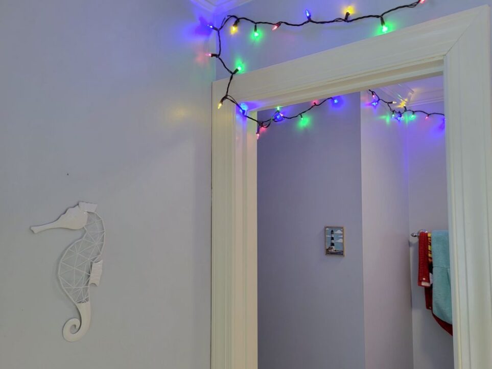 View of upstairs hallway bathroom standing inside sink area, facing the door opening that leads into the toilet/tub area. A lit strand of multi-coloured string lights is seen tacked along the ceiling of the sink area and snaking under the doorframe into the toilet/tub area where it continues to wind along the top of the ceiling there. On the wall to the left, in the sink area, hangs a simple white metal seahorse decoration. Ahead on the wall in the toilet nook hangs a small lighthouse decoration on the wall. Further ahead to the right is seen a towel rack (next to tub/shower) with children's towels hanging, partially out of sight.