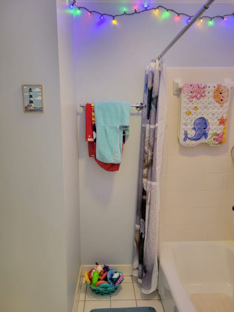 View of hallway bathroom back area with shower/tub. On back wall hangs small lighthouse picture and next to shower/tub is a towel rod with children's towels hanging. Along ceiling is lit strand of multi-coloured string lights. A colourful children's tub mat hangs on towel bar inside tub area.