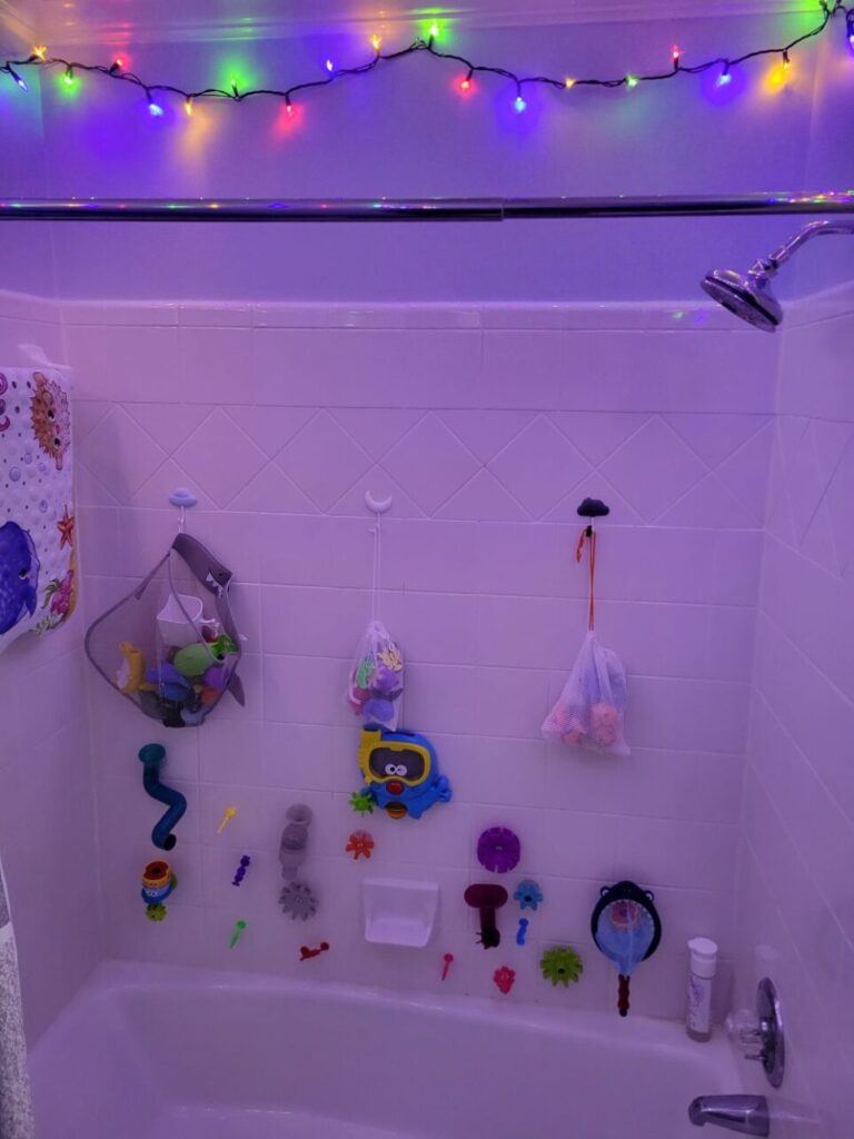 View of bathtub/shower in upstairs hallway bathroom. Across the top of the ceiling hangs a lit strand of multi-coloured string lights, creating a fun yet soothing glow to illuminate the tub area. Hanging on the white tile tub shower wall are various colourful children's bath toys.