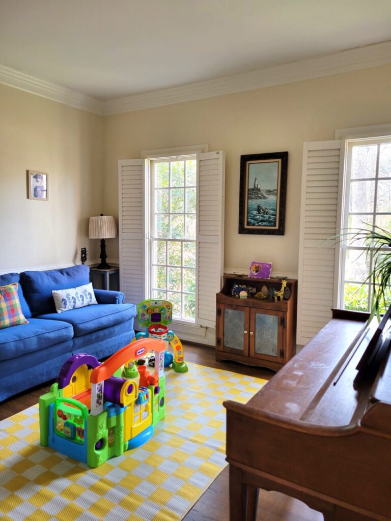 View of the front living room from doorway leading from the back living room (which connects the two living rooms). Alongside wall on right, most of the piano is seen. Ahead is the opposite wall with two windows facing the front yard. Between the two windows, a small antique table with son's artwork on top. Above it hangs a wood-framed painting of a coastal scene. In left corner is a small end table/plant stand with a lamp on top, next to it leans a guitar (mostly out of sight), and along left wall is a blue loveseat/sofa (partially out of sight). A framed photo hangs on wall above it. On floor in center is a yellow and white chequered area rug with a couple of large children's toys on top.