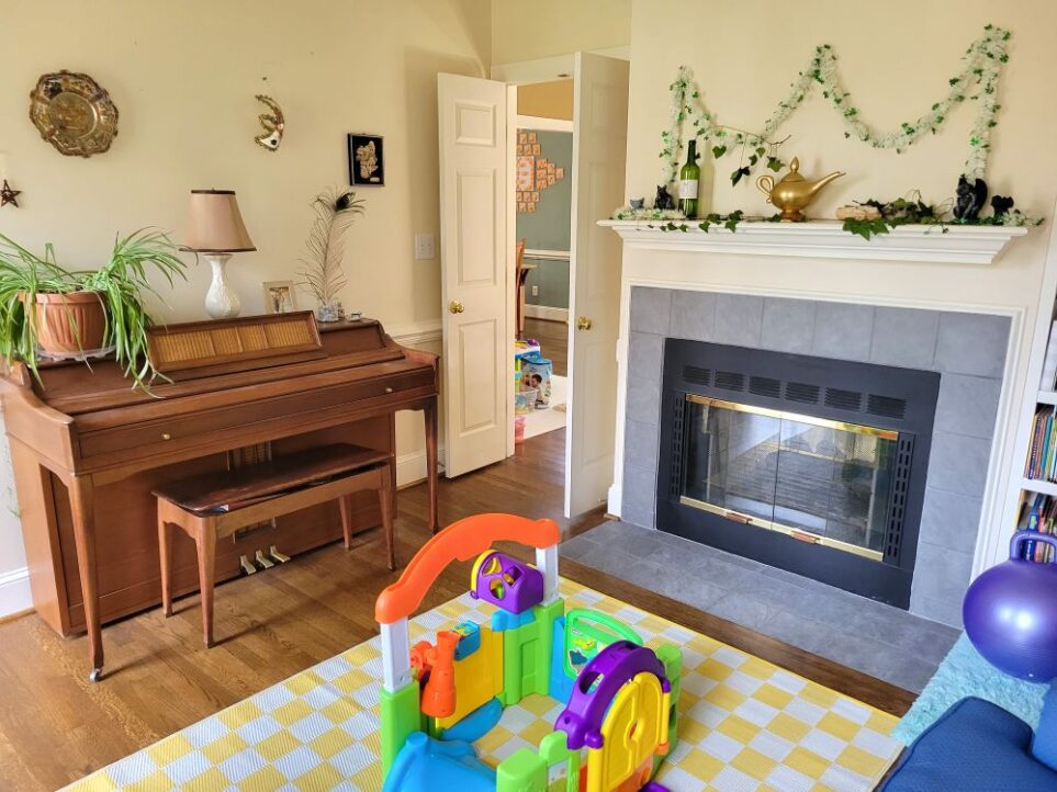 View of front LR facing the corner with the open French doors leading into the back LR. Along the left wall is a small wood piano with an airplane plant, lamp, framed photo, peacock feather and other decor featured on top. Above it on wall hang other decorative pieces - a decorative metal plate from Egypt, a Venetian mask, and carved decorations from Bethlehem. Along the wall to the right is the fireplace and mantel above it decorated with seasonal decorations - a St. Patrick's Day garland, ivy clippings, etc. Out of sight to the right of fireplace is built-in bookshelf. On floor in center of room is a yellow and white chequered area rug and children's toy on top.