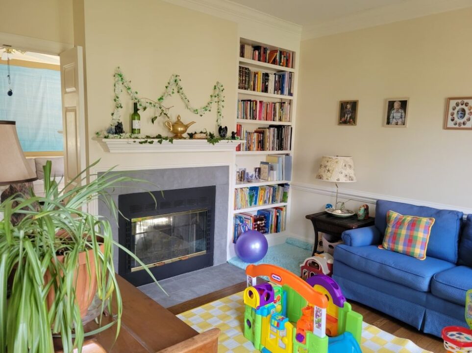 View of front LR as seen from its entrance from the foyer, facing the corner with the built-in bookcase. Along the wall to the right is a blue loveseat in partial view with a colourful chequered throw pillow. Flanking the left end of the loveseat is a small wicker end table with a coastal lamp on top. Below that on the floor to the left is an aqua shag rug in the reading nook. To its left built into the wall/corner is a bookcase filled with children's books on lower shelves and adult books up upper shelves. A large purple bouncy ball rests on rug against bookcase. To left of bookcase is fireplace and mantel. A few framed photos hang on right side wall. On floor is a yellow-and-white chequered area rug with children's toys on it (partially out of sight). To immediate left view in photo is part of the piano along left wall (out of sight) and lamp and airplane plant on top. A view of the back living room is seen through the open French doors in the left corner.