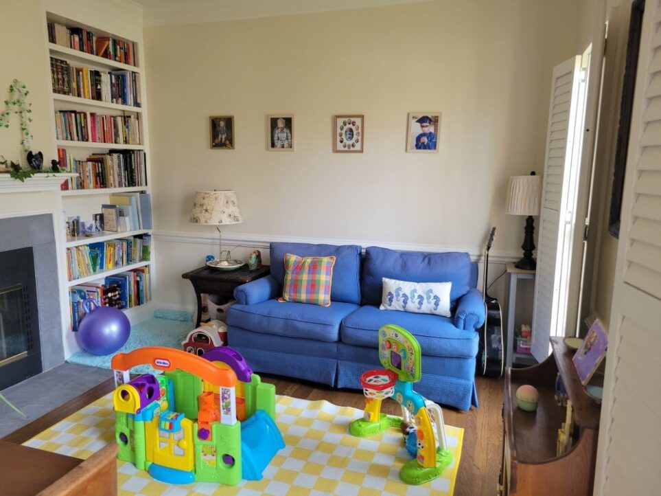 View of front living room as seen from its entrance from the foyer, facing the wall with a blue loveseat. In the left corner is a built-in bookcase and small aqua shag rug on floor between it and the sofa's end table with a lamp on top to create a cosy reading corner. To the left of the bookcase (and partially out of sight) along left wall is the fireplace and mantel. On the loveseat are a colourful chequered throw pillow and seahorse throw pillow. Above it on wall are four framed photos of kids. To its right stands a guitar in between it and a small plant stand/end table with a lamp on top in the right corner. Along the right wall are two windows with open plantation shutters and a small brown table in between. On floor is a yellow-and-white chequered area rug with children's toys on it.