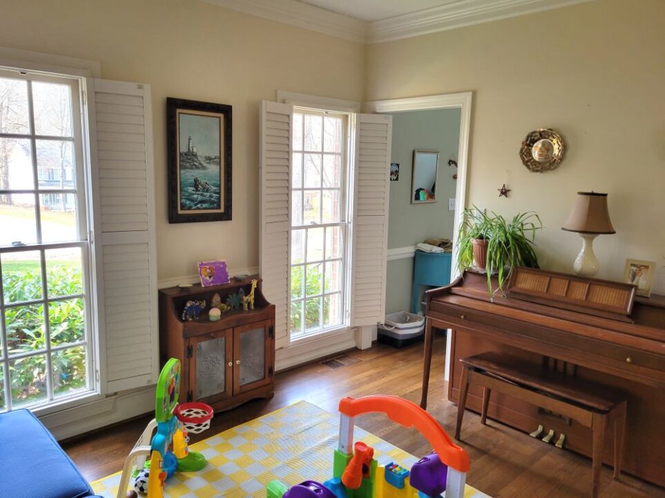 The front living room, distinguished as the one of two living rooms positioned in the front of the house, features two windows with plantation shutters and a small antique wood table in between featuring son's painted wooden figures. Above it hangs a wood-framed coastal painting. On the right side of the room along next wall is a small piano with a lamp on top, framed photo, and airplane plant. A couple of small decorations hang on the wall above it. A yellow and white chequered area rug is partially in view in center of floor along with a couple of colourful children's toys.