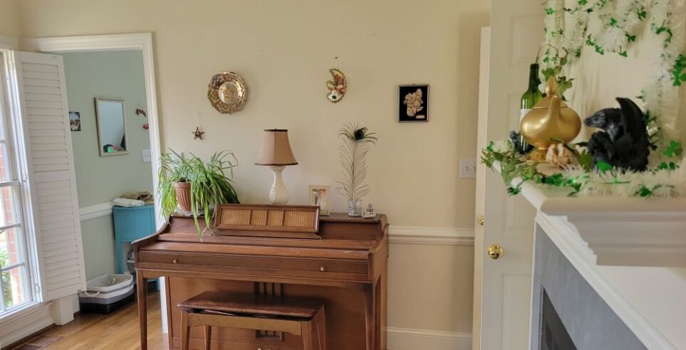 Front living room viewed from cosy corner facing piano wall: along right line of sight is mantel at eye level, with figurines, tinsel and ivy strung along it. Straight ahead against opposite wall is piano decorated with an upright peacock feather sticking out of a clear jar of round gray/translucent pebbles, a framed photo, shells, and an airplane plant. Decorations hang on wall above. Left of piano is opening that leads into foyer. Along wall left of that corner is (in partial sight) one of the two windows with open plantation shutters.