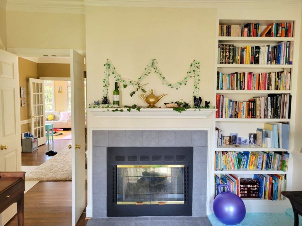 View of front LR facing fireplace wall. In left corner is double set of open French doors leading into the back LR, connecting the two rooms. (Barely in view along left wall the piano sticks out). Straight ahead is fireplace with black frame, glass doors with gold trim, and surrounding gray tiles with matching gray tile hearth flush with wood floor. Above fireplace is mantel decorated with various sentimental items - gold Aladdin's lamp teapot, gargoyle figurines (former favourites of son), local quartz stones, a tinsel garland strung along wall above in white with green shamrocks, a green wine bottle with ivy climbing out, and another vine of ivy strung along length of mantel. In left corner is a built-in bookcase sunken in wall with shelves up to nearly the ceiling. Bottom few have children's books as well as a few framed photos and trinkets. Top several shelves have adult books. On floor below bookcase is aqua shag rug in cosy reading corner. A large purple bouncy ball rests on it against bookshelves.