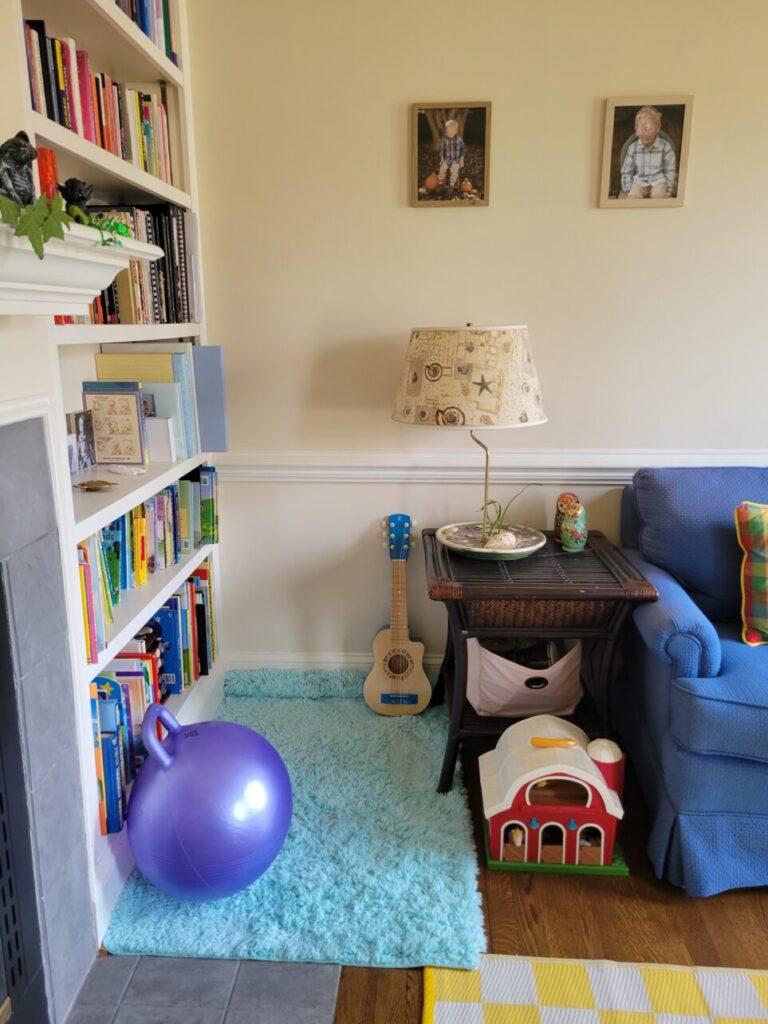 View of cosy corner in front living room, facing the end table next to the blue loveseat (mostly out of view). Atop end table is a costal lamp. Below it on shelf is a fabric bin with toy instruments. On floor in front of it is toy bar. On floor to left of it (in corner) is an aqua shag rug overtop a bath mat for added cushion. Standing along wall next to table is toy guitar. To left, beginning adjacent wall, is a built-in bookcase filled with children's books on bottom few shelves and adult books on upper shelves. A large purple bouncy ball rests on rug against bookcase. To left (and mostly out of sight) is fireplace and mantel. Two framed photos hang on wall above end of loveseat and end table. The corner of the yelllow-and-white chequered area rug is seen toward bottom right of photo (mostly out of sight)