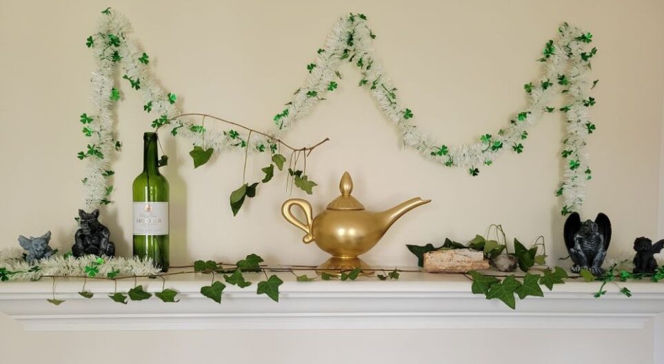 Close-up photo of the mantel in the front living room. Both living rooms share a double-sided fireplace, and each has a mantel which is decorated according to the season along with generally beloved keepsakes such as gold replica of Aladdin's lamp on mantel in this photo (which is also a functional teapot) and small gargoyle figurines which used to be older son's favourite toys. A few quartz rocks rest alongside ivy clippings and a St. Patrick's day white and green garland strings along wall above.