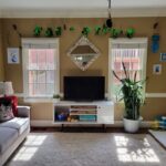 Back living room view of wall with two windows and white console table in between with TV screen on top and diamond-shaped mirror on wall above with a frame of seashells. Strung above the windows and mirror near ceiling is a garland of shiny green shamrocks for St. Patrick's Day. A blue evil eye keychain hangs from a black plastic strand of beads connected to the ceiling fan pull cord (out of view). In front of the right window is a large snake plant. To its right, a small white rug lays on the floor in the corner by the built-in bookcase with toys and a lamp on the lower shelves and seashell art decor on the top shelves (partially in view). On the wall right of the right window are two beachy framed photos, and on the wall to the left of the left window is a blue and purple print decoration. Right of the right window also hang bright blue shell chimes. A few toys are on the floor near the right corner. In the left corner is a small end table (out of sight) with lamp on top, and along the left wall next to it is the gray couch and above it the blue tapestry. Most of the floor is covered in a light-coloured high-pile rug.