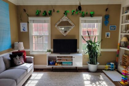 Back living room view of wall with two windows and white console table in between with TV screen on top and diamond-shaped mirror on wall above with a frame of seashells. Strung above the windows and mirror near ceiling is a garland of shiny green shamrocks for St. Patrick's Day. A blue evil eye keychain hangs from a black plastic strand of beads connected to the ceiling fan pull cord (out of view). In front of the right window is a large snake plant. To its right, a small white rug lays on the floor in the corner by the built-in bookcase with toys and a lamp on the lower shelves and seashell art decor on the top shelves (partially in view). On the wall right of the right window are two beachy framed photos, and on the wall to the left of the left window is a blue and purple print decoration. Right of the right window also hang bright blue shell chimes. A few toys are on the floor near the right corner. In the left corner is a small end table (out of sight) with lamp on top, and along the left wall next to it is the gray couch and above it the blue tapestry. Most of the floor is covered in a light-coloured high-pile rug.