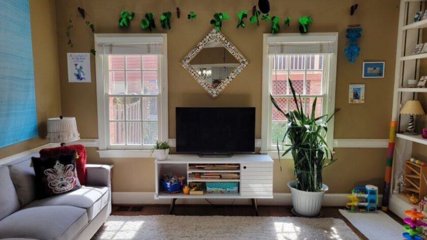 Back living room view of wall with two windows and white console table in between with TV screen on top and diamond-shaped mirror on wall above with a frame of seashells. Strung above the windows and mirror near ceiling is a garland of shiny green shamrocks for St. Patrick's Day. A blue evil eye keychain hangs from a black plastic strand of beads connected to the ceiling fan pull cord (out of view). In front of the right window is a large snake plant. To its right, a small white rug lays on the floor in the corner by the built-in bookcase with toys and a lamp on the lower shelves and seashell art decor on the top shelves (partially in view). On the wall right of the right window are two beachy framed photos, and on the wall to the left of the left window is a blue and purple print decoration. Right of the right window also hang bright blue shell chimes. A few toys are on the floor near the right corner. In the left corner is a small end table (out of sight) with lamp on top, and along the left wall next to it is the gray couch and above it the blue tapestry. Most of the floor is covered in a light-coloured high-pile rug.