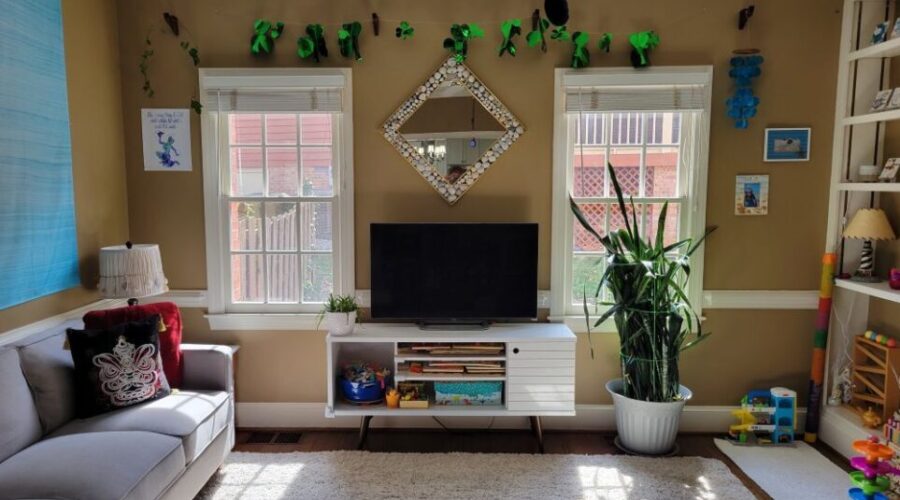 Back living room view of wall with two windows and white console table in between with TV screen on top and diamond-shaped mirror on wall above with a frame of seashells. Strung above the windows and mirror near ceiling is a garland of shiny green shamrocks for St. Patrick's Day. A blue evil eye keychain hangs from a black plastic strand of beads connected to the ceiling fan pull cord (out of view). In front of the right window is a large snake plant. To its right, a small white rug lays on the floor in the corner by the built-in bookcase with toys and a lamp on the lower shelves and seashell art decor on the top shelves (partially in view). On the wall right of the right window are two beachy framed photos, and on the wall to the left of the left window is a blue and purple print decoration. Right of the right window also hang bright blue shell chimes. A few toys are on the floor near the right corner. In the left corner is a small end table (out of sight) with lamp on top, and along the left wall next to it is the gray couch and above it the blue tapestry. Most of the floor is covered in a light-coloured high-pile rug.