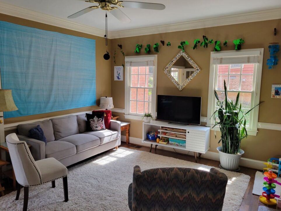 Back living room viewed from doorway connecting both living rooms and facing the back corner by the windows and couch. Just in view in front is back of an armchair, and to left is an armless upholstered chair. Most of the floor is covered in light high-pile rug. The gray couch lines the left wall under the bright blue wall hanging/tapestry. On next wall toward right are two windows and white console table in between with TV screen on top and diamond-shaped mirror above with a frame of seashells. Strung above the windows and mirror near ceiling is a garland of shiny green shamrocks for St. Patrick's Day. A ceiling fan hangs from ceiling just in view and a strand of black plastic beads hangs from it to reach the cord, with a blue evil eye affixed to the end. In front of the right window is a large snake plant. In corner by couch and window is end table with lamp. To left of couch (partially out of view, and by armless chair) is another small end table and floor lamp.