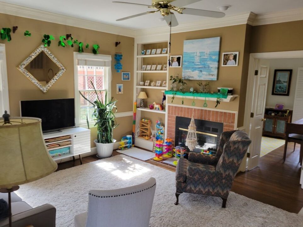 View of back living room facing the fireplace corner with built-in bookcase. Toward the right (mostly in view) are the open French doors leading into the front living room. Left of that doorway is the fireplace (which is a double fireplace and opens on the other side into the front living room) and mantel above it, decorated in a green scarf for St. Patrick's Day along with ivy cuttings and other green cuttings stuck out of clear glass vases and shamrock decorations hanging over edge. Above the mantel, on the wall, hangs a large print of a painting in light blues and white of two sailboats on the ocean. On either side of the picture are two framed photographs. On the slightly elevated brick hearth in front of fireplace are several colourful children's toys. Left of the fireplace in the back corner is a built-in bookcase with toys on the lower shelves and decorations on the top. Left of that is the south-facing wall with two windows (the left one barely in view) and a large snake plant in front of the right window. Two upholstered chairs anchor both sides of the area rug, one near the hearth and one near the couch (the couch as well as a floor lamp next to it are barely in view, lining the wall to the left).