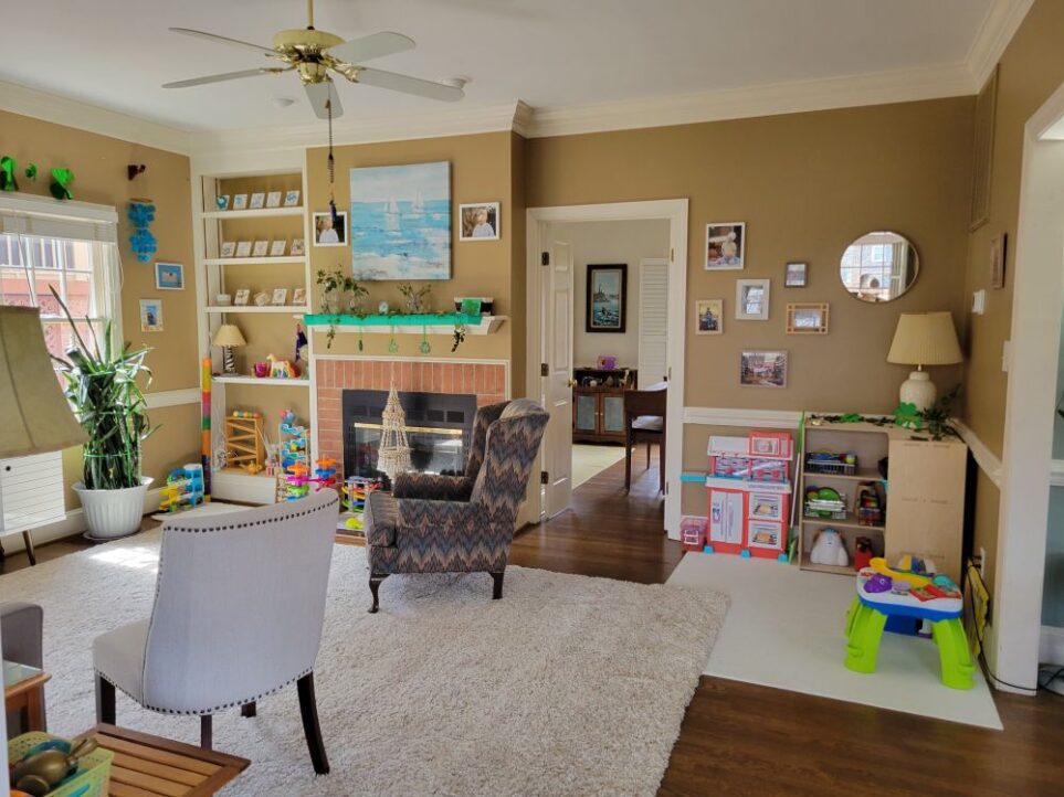 View of back living room from the open doorway leading into the sunroom, facing the fireplace wall and open door to front living room. In the right corner, between the open French doors leading into the front living room straight ahead and the opening leading into the kitchen on the right (mostly out of sight), is a toy nook/play space with a Montessori corner toy shelf positioned in the corner. Atop it is a lamp as well as St. Patrick's Day decor and ivy clippings. Beside it are a toy kitchen on one side and a toddler table on the other, with several toys filling the shelves inside. A thin medium-sized area rug lays across the area in that corner as well (partially under a corner of the larger area rug). To the left of the open door to the front living room is the fireplace (which is a double fireplace shared by both living rooms and which also opens on the other side into the front living room) and mantel above it, decorated in a green scarf for St. Patrick's Day along with ivy cuttings and other green cuttings stuck out of clear glass vases and shamrock decorations hanging over edge. Above the mantel, on the wall, hangs a large print of a painting in light blues and white of two sailboats on the ocean. On either side of the picture are two framed photographs. On the slightly elevated brick hearth in front of fireplace are several colourful children's toys. Left of the fireplace in the back corner is a built-in bookcase with toys on the lower shelves and decorations on the top. Left of that is the south-facing wall with windows (one is partially in view) and a large snake plant in front of window. Two upholstered chairs anchor both sides of the area rug, one near the hearth and one near the couch.
