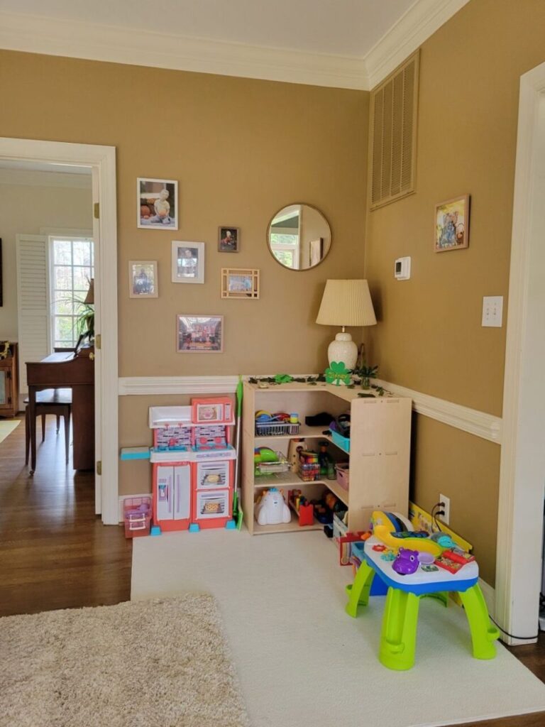 In a corner nook of the back living room, between the open French doors leading into the front living room on the left side and the wide opening leading into the kitchen on the right side, is a toy nook/play space with a Montessori corner toy shelf positioned in the corner. Atop it is a lamp as well as St. Patrick's Day decor and ivy clippings. Beside it are a toy kitchen on one side and a toddler table on the other, with several toys filling the shelves inside. A thin medium-sized area rug lays across the area in that corner as well (partially under a corner of the larger area rug).