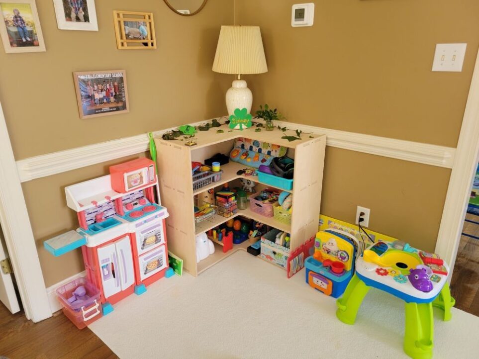 The Montessori corner toy shelf is seen in the back living room corner between the opening to the kitchen (out of sight, to right) and opening to the front living room (out of sight, to left). Atop it is a lamp as well as St. Patrick's Day decor and ivy clippings. Beside it are a toy kitchen on one side and a toddler table on the other, with several toys filling the shelves inside. A thin medium-sized area rug lays across the area in that corner as well (partially under a corner of the larger area rug). The toy space is bright, playful and inviting, as well as neatly tucked out of the main walking area in the living room.
