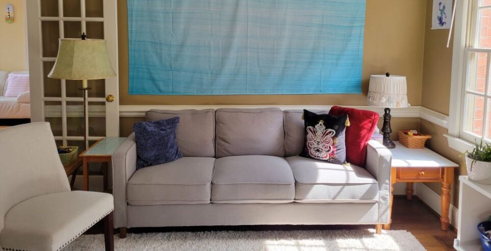 The new gray couch (a basic/standard three-seater couch) positioned along the wall in the back LR, with a few throw pillows on it and two small end tables and lamps on either side. To its right is the south-facing wall with a window and plant and diagonal to its left is a chair. Beyond the left-side end table and floor lamp is the open French door leading into the sunroom (mostly out of sight)