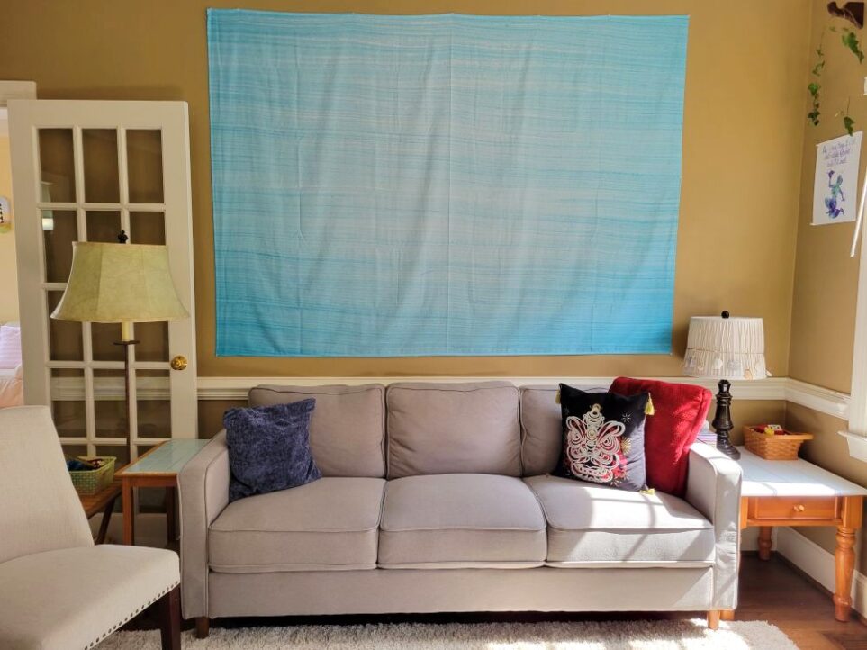 A large blue wall tapestry (a light thin fabric) hangs above the gray couch in the back living room to cover a large expanse of the wall, in order to minimize the view of its paint colour.
