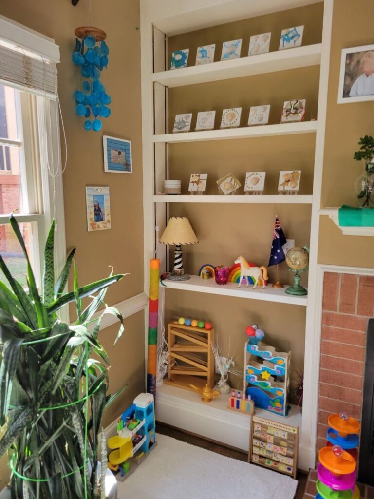 The built-in corner bookcase in back living room features seashell tile art displayed on top three shelves and colourful children's toys and a small lighthouse lamp on the lower two shelves. Bright blue shell chimes hang from ceiling to the left, next to the window. A very large snake plant is in front of the window, to the left. To the right (and mostly out of sight) is the fireplace and mantel. A few other toys are tastefully stationed on the floor in front of the bookcase, with a small white rug (overtop an old bath mat for cushioning) on the floor in front of it as a mini play space.
