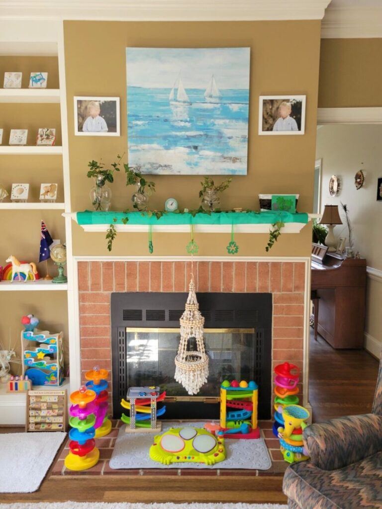 View of fireplace in back living room (which is a double fireplace shared by both living rooms and also opens on the other side into front living room) and mantel above it, decorated in a green scarf for St. Patrick's Day along with ivy cuttings and other green cuttings stuck out of clear glass vases and shamrock decorations hanging over edge. Above mantel, on wall, hangs a large print of a painting in light blues and white of two sailboats on the ocean. On either side of the picture are two framed photographs. On the slightly elevated brick hearth in front of fireplace are several colourful children's toys which, despite being of an entirely different decorative aesthetic from the rest of the room, are neatly arranged in good order and add a very cheerful vibe to the room.