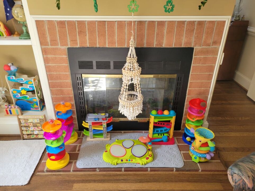 The fireplace in the back living room (which is a double fireplace shared by both living rooms and also opens on the other side into front living room) features a slightly raised brick hearth to match the brick fireplace. Covering the hearth is a gray bathmat in the center with several colourful children's toys arranged on top of it and on either side - a car racer ramp, similar ball ramp, a few spiral ball toys, etc. St. Patrick's Day shamrock decorations hang from edge of the mantel at the top of the photo. Partially in view to right is opening into front living room, and partially in view to left is bottom two shelves of built-in bookcase with more toys arranged on them.