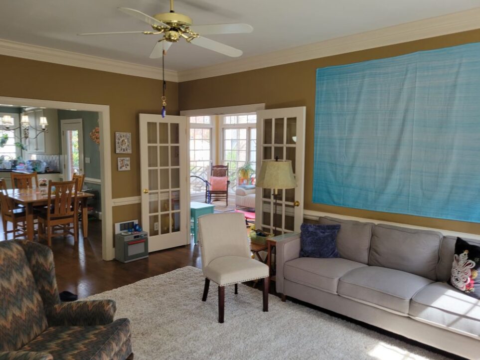 View of back living room from the fireplace hearth, facing the corner with the open French doors into the sunroom. To the right along the wall is the gray couch and above it the blue wall tapestry. Next to the couch on left side are a couple of small end tables and floor lamp. On next wall to left of open door into sunroom is a small toy dishwasher and then the wide opening into the breakfast area of the kitchen with kitchen table. To left side of photo is an armchair (partially in sight) and straight ahead, next to couch (in front of end table), is an armless upholstered chair. On most of the floor of the back living room is a large light-coloured high-pile area rug. Above hangs a ceiling fan and, from it, a blue evil eye keychain affixed to a black plastic strand of beads hanging from the fan pull cord. A couple of framed photos hang on the wall right of the breakfast area doorway, above the toy dishwasher. A couple of throw pillows are on the gray couch. More furniture and plants can be seen straight ahead in back corner in sunroom through the open doorway.