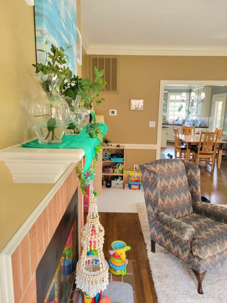 View of back LR standing in corner by built-in bookcase and looking along edge of mantel (at eye level) straight to back corner with toy shelf. Along mantel is a bright green scarf for St. Patrick's Day and several clear glass vases filled with ivy clippings. Hanging from mantel is a large cowrie shell chandelier, which hangs amidst toys on hearth below. To right is an armchair on the edge of the area rug, and straight ahead in back corner is toy shelf with toys. To the right of it is the opening into the breakfast area of the kitchen where kitchen table and chairs are in view.