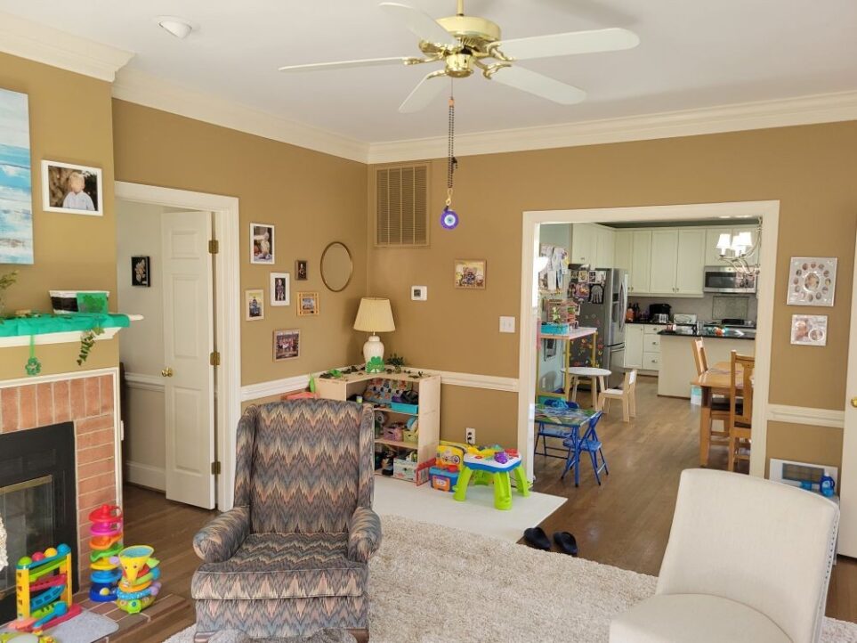 Back living room view from the couch, facing the back corner with the toy shelf and opening into the kitchen. To left (mostly out of sight) is fireplace with toys on hearth and St. Patrick's Day decor on mantel. On area rug in center of shot are two chairs anchoring either side of the rug. Beyond chair on left and back in corner is Montessori toy shelf with lamp and toys. Left of it (and to the right of the fireplace) is the entry into the front living room. To the right of the toy corner is the wide opening into the kitchen breakfast area with toddler tables and kitchen table in view as well as functional kitchen beyond it, a nice open space for the whole family to be together.