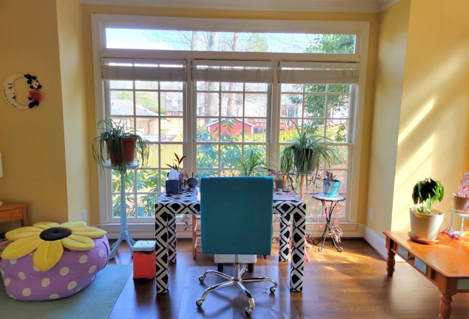 View of study directly facing east wall with three windows. Several plants on plant stands are directly in front of the windows. In front of them is black-and-white patterned upholstered desk and teal swivel chair, with a couple more plants on top along with other items like tissue box and blue ceramic seashell pen holder. To right partially seen is coffee table with more plants on top (facing south-window, out of sight) and to left is beanbag chair filled with pillows and sunflower pillow on top, over small blue corner rug.