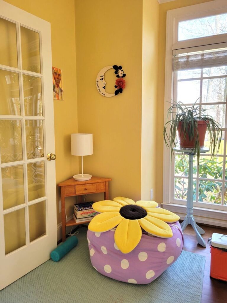 View of cosy corner in study: to left is open French door, on right is the first window and an airplane plant on a blue-painted plant stand in front of it. In corner is a small triangular wood table with a white lamp on top, and several books stacked on the lower shelf. In front of it is a large periwinkle with white polka-dot beanbag filled with pillows and a soft flat sunflower pillow on top. On the wall above the lamp hangs a portrait painting on the left side and a wood crescent moon with a face and flowers on the other side