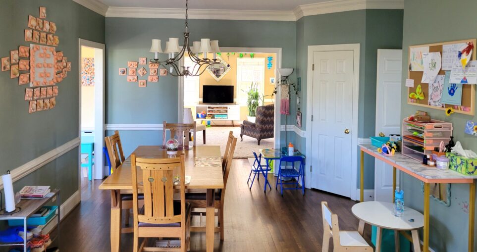 Breakfast area of kitchen facing the opening into back living room (from standing in front of the counter island). To left center of space is kitchen table and chairs. To right is short wall with console table and round white toddler table. Beyond it is opening into foyer, and left of that is the closed pantry door. To left of pantry door is second toddler table where kindergartener prefers to sit, along with a floor lamp in corner with macrame design hanging from switch and some artwork on the small wall space above table. To immediate left is wide opening into the back living room. Left of it is small span of wall with seashell artwork on it (and a toy school bus on floor in small corner nook, mostly out of sight), and immediately to left along next wall is opening into sunroom. Seashell art hangs on the left wall, and below in near left corner is shoe rack (mostly in view).