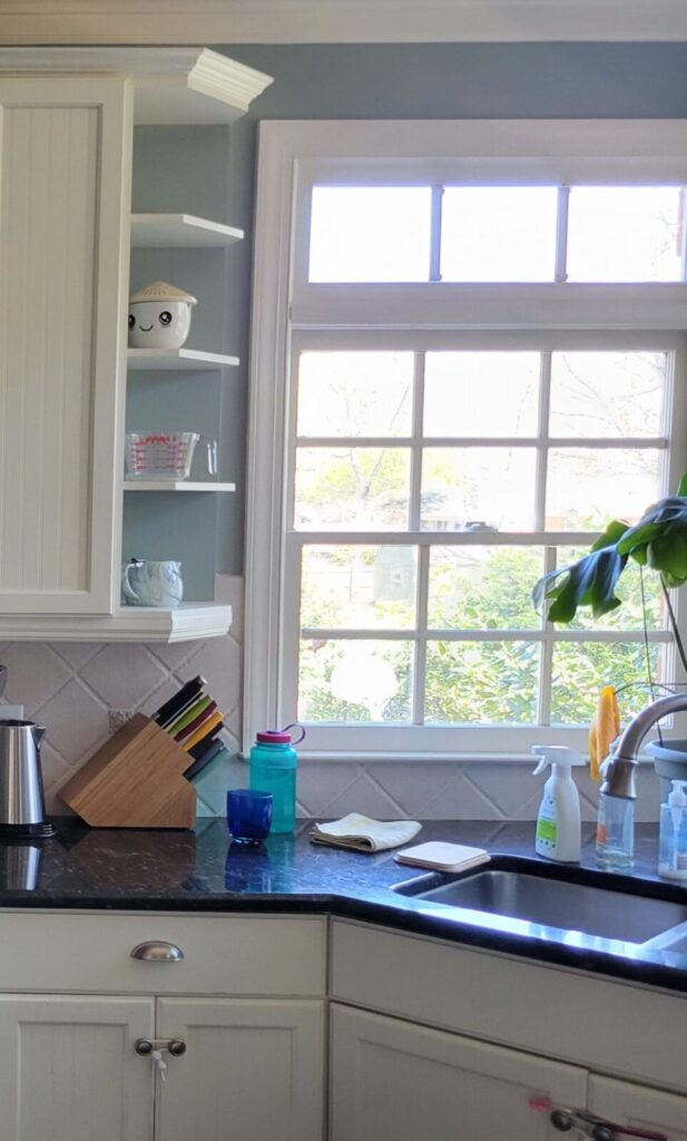 Partial view of kitchen sink, window behind it, cabinetry below it, and cabinetry above and to left, with a few open shelves and dishes on them