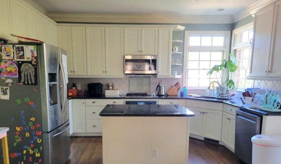 The functional kitchen, seen in photo, is the command center of the kitchen and dining room. To the left is the refrigerator with photos and magnets stuck to the side. In the center is a countertop island. Against opposite wall is countertop and cabinets with oven, stove and microwave in center. In far right corner is kitchen window and wink. To right is dishwasher and, against end of counter, trash bin.