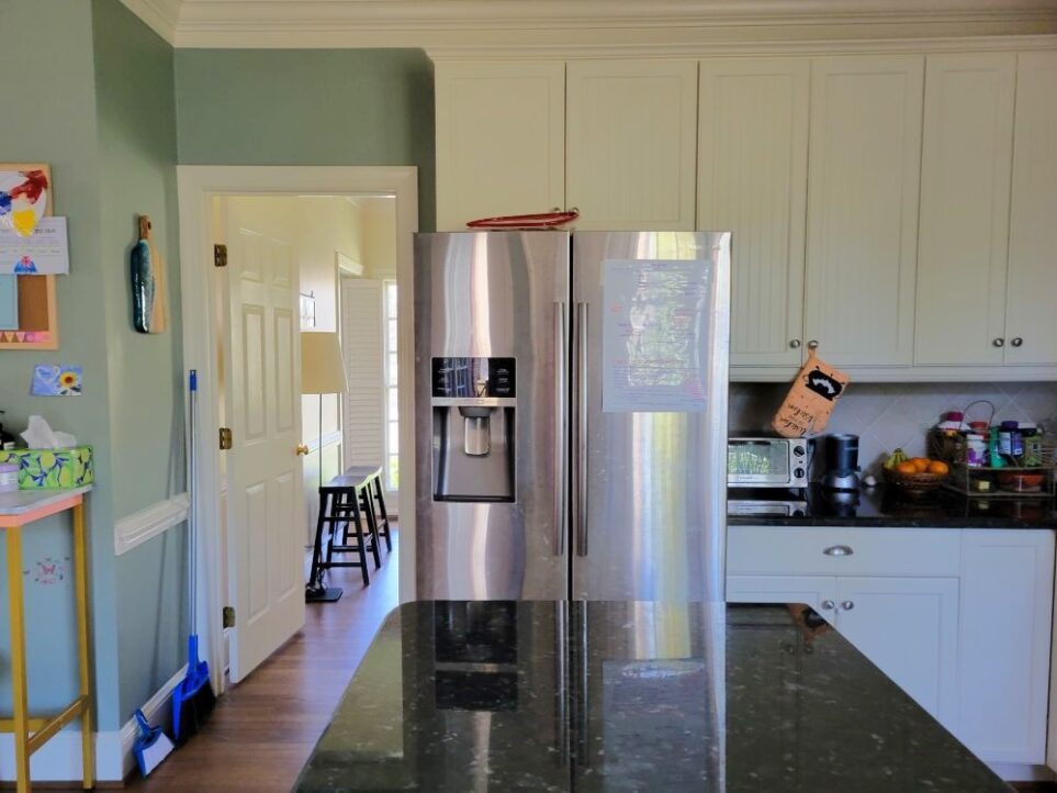 View of part of the kitchen and dining room, from standing on the dishwasher end of the countertop island. Straight ahead is refrigerator, and to the right of it, beginning of countertop and cabinets with toaster oven, NutriBullet, produce, vitamins, and other things that lack pantry or accessible cabinet space. Above refrigerator is empty cabinet space that is too high to reach (and completely inaccessible, even to clean, because of inability to reach inside while the door is open, from standing either on the counter beside it or in front of the frig below it, because the refrigerator - which also doesn't work right - is so large). Left of the refrigerator is the open dining room door, showing a floor lamp, two black stools, and (barely in view) a window on the front wall. Left of open dining room door is a broom leaning in corner and beginning of console table and bulletin board on wall above it (mostly out of sight).