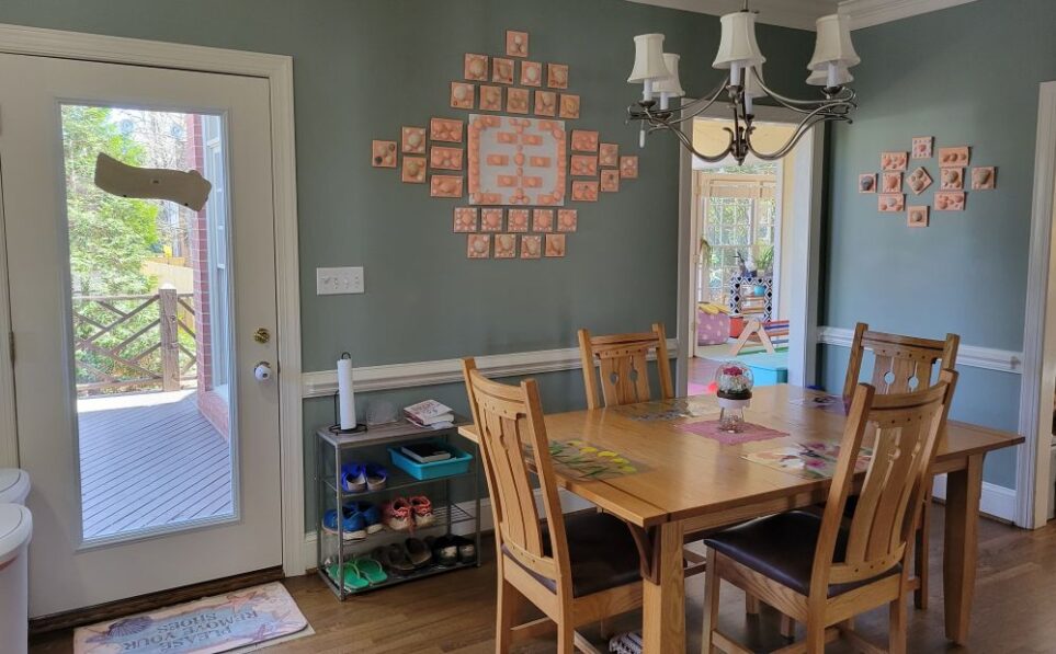 Breakfast area of kitchen viewing wall with back door and entrance into sunroom in back right corner. To the right of the back door is a small shoe rack with a few pairs of shoes and, on the top shelf, a paper towel roll/holder and a couple of books. In teal bin on second shelf are more books and magazines. Seashell tile art hangs in a gallery on wall above, as well as on wall to the right of the opening into the sunroom. The kitchen table and chairs are in the center of the space, and above them hangs a silver candelabra-style chandelier with small white lampshades.