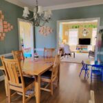 The breakfast area viewed from the functional kitchen and dining room doorway. The wood table and chairs are in the left center, under a candelabra-style silver chandelier with small white lampshades. On wall to left hangs seashell artwork collage. Partially in view on floor to left is shoe rack. Barely in view to front right is toddler table. Ahead to right (just left of partially-seen closed pantry door) is another toddler table (where kindergartener likes to sit, instead of the big table) and, behind it, a floor lamp. In front of it (and spanning wall toward left) is wide opening into back living room. Beyond remaining wall space (where more seashell artwork hangs) to left corner is opening into sunroom.