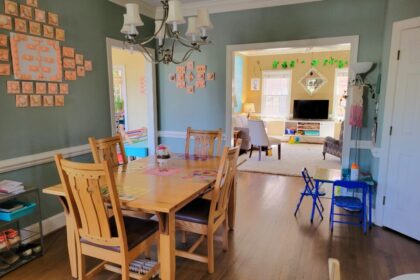 The breakfast area viewed from the functional kitchen and dining room doorway. The wood table and chairs are in the left center, under a candelabra-style silver chandelier with small white lampshades. On wall to left hangs seashell artwork collage. Partially in view on floor to left is shoe rack. Barely in view to front right is toddler table. Ahead to right (just left of partially-seen closed pantry door) is another toddler table (where kindergartener likes to sit, instead of the big table) and, behind it, a floor lamp. In front of it (and spanning wall toward left) is wide opening into back living room. Beyond remaining wall space (where more seashell artwork hangs) to left corner is opening into sunroom.