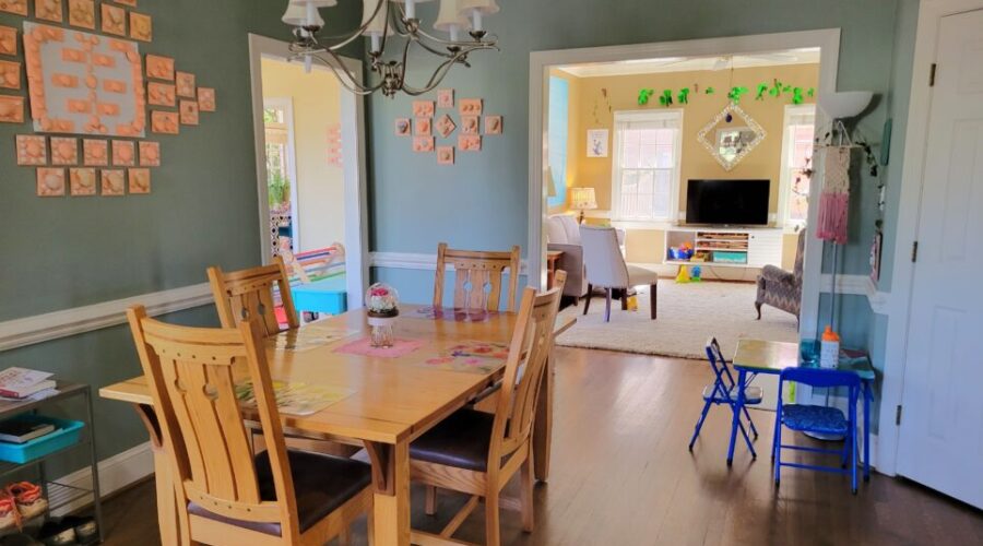 The breakfast area viewed from the functional kitchen and dining room doorway. The wood table and chairs are in the left center, under a candelabra-style silver chandelier with small white lampshades. On wall to left hangs seashell artwork collage. Partially in view on floor to left is shoe rack. Barely in view to front right is toddler table. Ahead to right (just left of partially-seen closed pantry door) is another toddler table (where kindergartener likes to sit, instead of the big table) and, behind it, a floor lamp. In front of it (and spanning wall toward left) is wide opening into back living room. Beyond remaining wall space (where more seashell artwork hangs) to left corner is opening into sunroom.