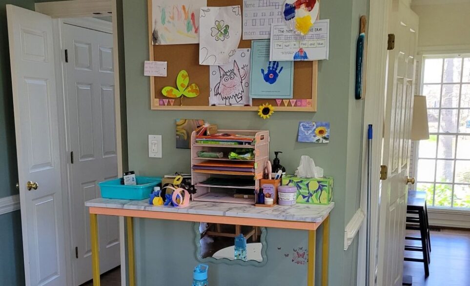 Close-up view of top of console table and bulletin board hung above it in breakfast area of kitchen. The top is faux marble and the base (under the top 'lip') is covered in a ribbon of sparkly orange peel-and-stick wallpaper. Several children's artworks are hung on the bulletin board, creating a colourful collage and visual happiness.