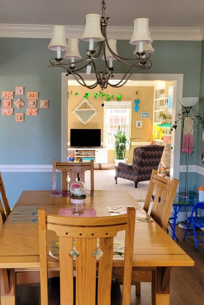 Kitchen table and chairs situated below silver-coloured candelabra-style chandelier with small white lampshades. Replacing the small lightbulbs with LEDs now allows a much brighter lighting capacity; still, there is an extra floor lamp seen in the back right corner (by the square toddler table) for maximum lighting when/as needed.