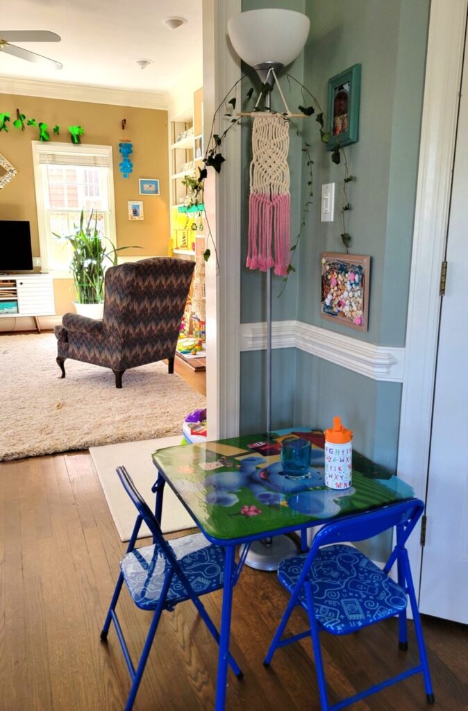 Corner of kitchen breakfast area (nook between pantry on right and wall opening to back living room on left) with square toddler table and floor lamp. The floor lamp offers a nice light for this corner of the room and also allows more pleasant decor such as the tan and pink macrame decoration ($2 from a thrift store) and ivy tendrils from the yard, alongside framed artwork and photo on the small section of wall.