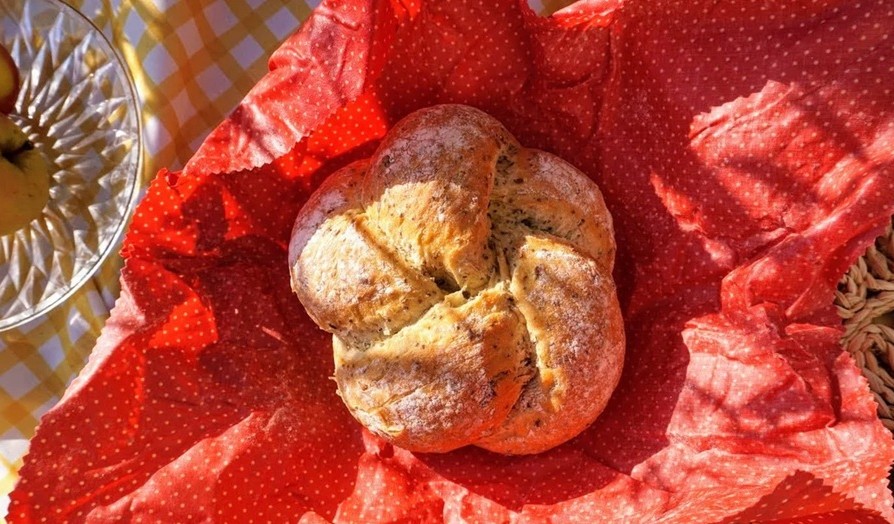 A homemade loaf of bread rests on top of a red beeswax bread wrap, ready to be wrapped up in it